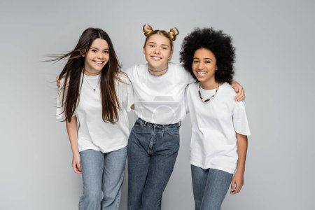 Smiling blonde teen girl in white t-shirt and jeans hugging multiethnic girlfriends and looking at camera on grey background, adolescence models and generation z concept