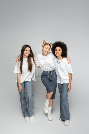 Full length of joyful blonde teen girl looking at camera while hugging multiethnic friends in white t-shirts and jeans and standing on grey background, adolescence models and generation z concept