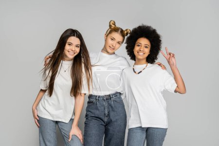 Trendy blonde teen girl in jeans and white t-shirt hugging multiethnic girlfriends and posing together isolated on grey, adolescence models and generation z concept, friendship and companionship