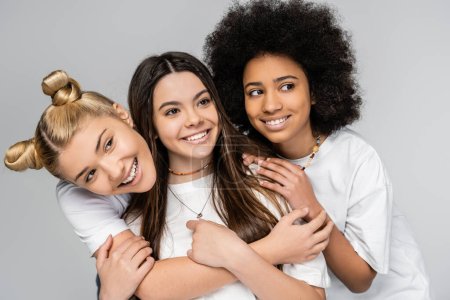 Foto de Retrato de adolescentes alegres y multiétnicos en camisetas blancas abrazando y mirando hacia otro lado mientras posan aislados sobre niñas grises, adolescentes y generación z concepto, amistad y compañerismo - Imagen libre de derechos