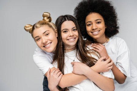 Photo for Portrait of cheerful and multiethnic teenage girls in white t-shirts hugging each other and looking at camera while standing isolated on grey, teenage friends having fun together - Royalty Free Image