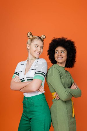 Lächelnde und multiethnische Teenager-Freundinnen in trendigen Outfits, die sich die Arme überkreuzen und Rücken an Rücken stehen, während sie isoliert auf orangefarbenem, trendigem Generation-Z-Konzept, Freundschaft und Kameradschaft posieren.