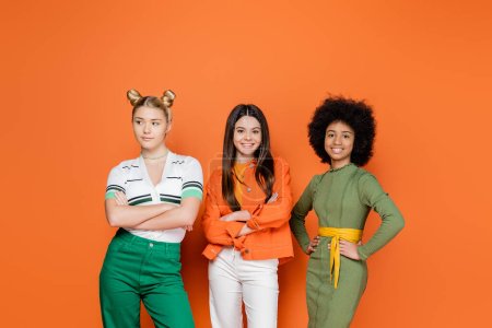 Pozytywne i międzyrasowe nastoletnie przyjaciółki w modnych strojach krzyżujących ramiona, stojące i pozujące razem na pomarańczowym tle, modne pokolenie z koncepcją, przyjaźń i towarzystwo