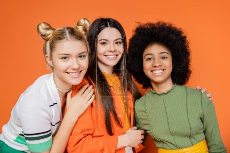 Retrato de chicas adolescentes de moda y multiétnicas en trajes de moda y maquillaje abrazando y mirando a la cámara mientras posan sobre fondo naranja, concepto de generación z de moda