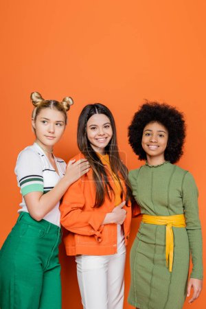 Lächelndes brünettes Teenie-Mädchen in stylischem Outfit, das in die Kamera neben multiethnischen Freundinnen blickt, während es gemeinsam auf orangefarbenem Hintergrund posiert, trendiges Generation-Z-Konzept, Freundschaft und Kameradschaft