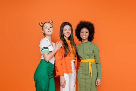 Téléchargez les photos : Groupe de modèles adolescents interracial en tenue élégante posant et souriant à la caméra tout en se tenant ensemble sur fond orange, concept tendance de génération z, amitié et compagnie - en image libre de droit