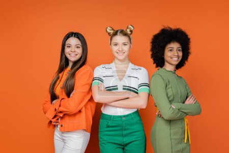 Selbstbewusste und modische multiethnische Teenager, die die Arme verschränken, während sie stehen und gemeinsam posieren auf orangefarbenem Hintergrund, trendiges Generation-Z-Konzept, Freundschaft und Kameradschaft