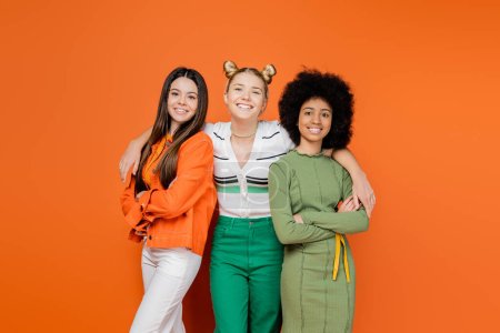 Cheerful blonde teenage girl hugging fashionable multiethnic girlfriends in trendy outfits crossing arms and looking at camera on orange background, trendy generation z concept