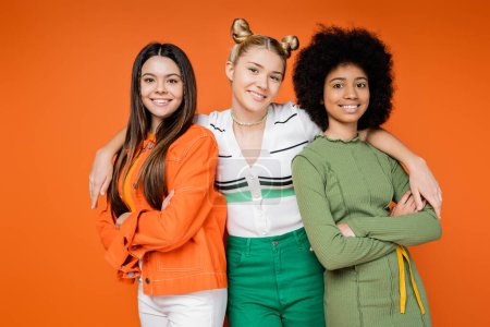 Portret radosnych i międzyrasowych nastoletnich dziewczyn w modnych strojach krzyżujących ramiona i patrzących w kamerę odizolowaną od pomarańczowej, kulturowej różnorodności i pokoleniowej koncepcji mody z