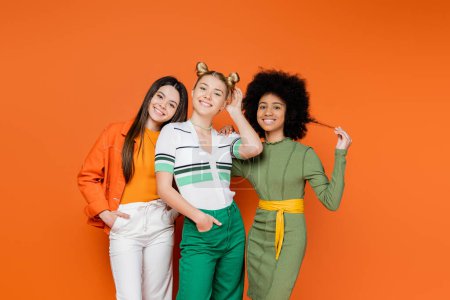 Téléchargez les photos : Copines multiethniques et adolescentes dans des tenues à la mode avec maquillage posant ensemble et regardant la caméra sur fond orange, la diversité culturelle et la génération z concept de mode, amitié - en image libre de droit