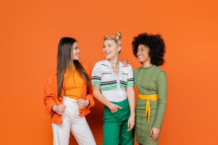 Fröhliche und multiethnische Freundinnen in trendigen Outfits reden und schauen einander an, während sie gemeinsam auf orangefarbenem Hintergrund stehen, kulturelle Vielfalt und generationenübergreifendes Modekonzept