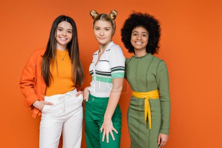 Stylish multiethnic teenage girlfriends in trendy outfits posing together and looking at camera while standing on orange background, cultural diversity and generation z fashion concept