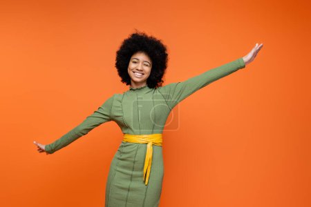 Cheerful and teen african american girl in stylish green dress and bold makeup winking and looking at camera while standing isolated on orange, teenage fashion and generation z concept 