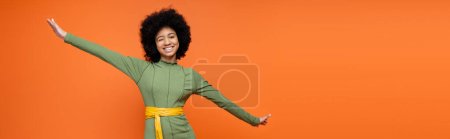 Foto de Sonriente chica adolescente afroamericana con maquillaje audaz con vestido verde y guiño a la cámara aislada en naranja, moda adolescente y concepto de generación z, pancarta con espacio para copiar - Imagen libre de derechos