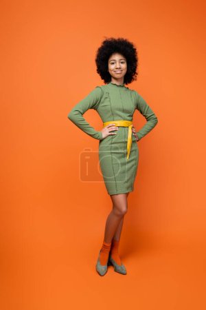 Full length of smiling and fashionable african american teen girl with bold makeup posing in green dress while standing on orange background, teenage fashion and generation z concept  