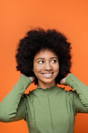 Portrait of joyful teenage african american girl with bold makeup and green dress touching hair and looking away isolated on orange, teenage fashion and generation z concept 