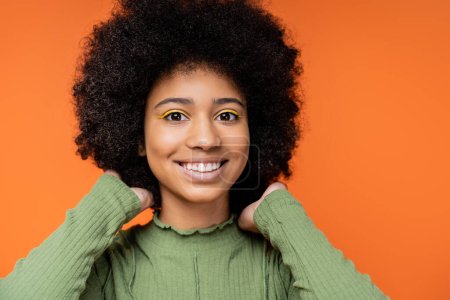Portrait of smiling and teenage african american girl with bold makeup in green dress touching hair and looking at camera isolated on orange, teenage fashion and generation z concept 