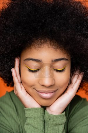 Portrait of smiling and trendy teen african american girl with bright makeup touching cheeks and posing isolated on orange, youth culture and generation z concept 