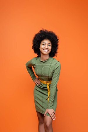 Trendiges und positives afrikanisch-amerikanisches Teenager-Mädchen mit hellem Make-up in grünem Kleid und Händchenhalten an der Hüfte isoliert auf orange, Jugendkultur und Generation-Z-Konzept 