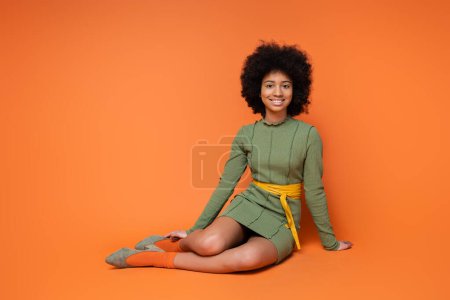 Foto de Chica afroamericana alegre y adolescente en vestido verde de moda sonriendo a la cámara mientras se sienta y posa sobre fondo naranja, cultura juvenil y concepto de generación z - Imagen libre de derechos