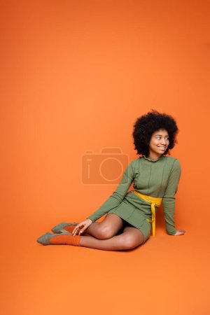 Positive und stilvolle afrikanisch-amerikanische Teenagerin in grünem Kleid, die wegschaut, während sie sitzt und auf orangefarbenem Hintergrund posiert, Jugendkultur und Generation-Z-Konzept 