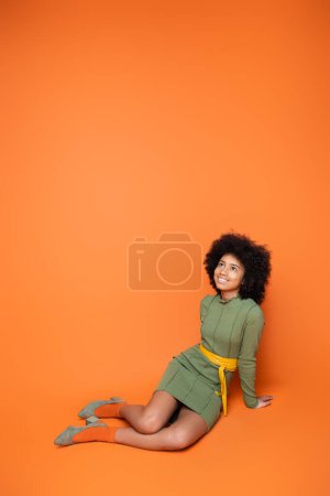 Foto de Longitud completa de adolescente afroamericano alegre y de moda en vestido verde mirando hacia arriba mientras está sentado y posando sobre fondo naranja, cultura juvenil y concepto de generación z - Imagen libre de derechos