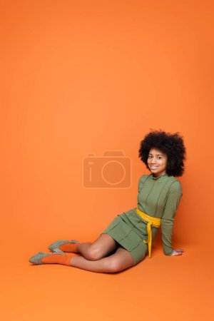Pleine longueur d'adolescent afro-américain positif avec un maquillage audacieux et une robe verte souriant à la caméra tout en étant assis sur fond orange, la culture des jeunes et la génération z concept 