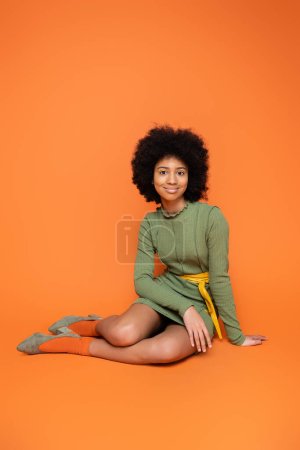 Full length of teen african american model with bold makeup wearing green dress while sitting and looking at camera on orange background, youth culture and generation z concept  
