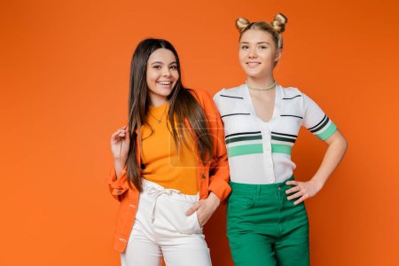 Positive brunette teenage model in casual outfit posing and standing with stylish blonde girlfriend together on orange background, fashionable girls with sense of style, friendship and bonding