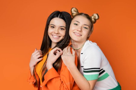 Portrait of smiling blonde teenage girl with colorful makeup hugging stylish brunette girlfriend and looking at camera isolated on orange, fashionable girls with sense of style, friendship 