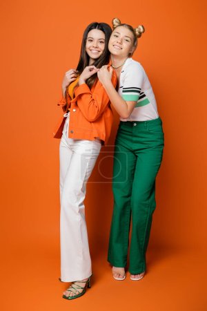 Volle Länge der blonden trendigen Teenager-Mädchen in lässiger Kleidung, die in die Kamera schaut, während sie brünette Freundin umarmt und auf orangefarbenem Hintergrund steht, modische Mädchen mit Sinn für Stil
