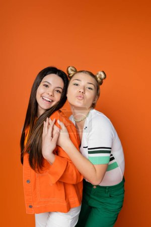 Portrait of stylish blonde teenage girl with bold makeup blowing air kiss at camera and hugging cheerful brunette girlfriend isolated on orange, fashionable girls with sense of style