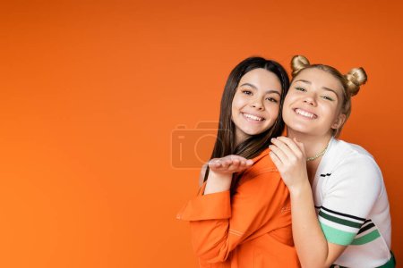 Fröhliche brünette Teenager-Mädchen mit hellem Make-up bläst Luftkuss in die Kamera und steht in der Nähe trendige blonde Freundin isoliert auf orangefarbenen, modischen Mädchen mit Sinn für Stil