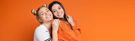 Positive and trendy blonde teenage model with bright makeup hugging brunette girlfriend and looking at camera isolated on orange, fashionable girls with sense of style, banner with copy space