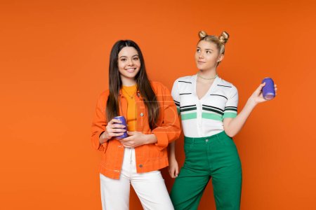 Smiling brunette teenage girl in trendy outfit holding drink in tin can and looking at camera near blonde girlfriend on orange background, fashionable girls with sense of style