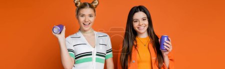 Photo for Joyful and stylish blonde and brunette teenage models looking at camera while holding drink in tin cans on orange background, fashionable girls with sense of style, banner - Royalty Free Image