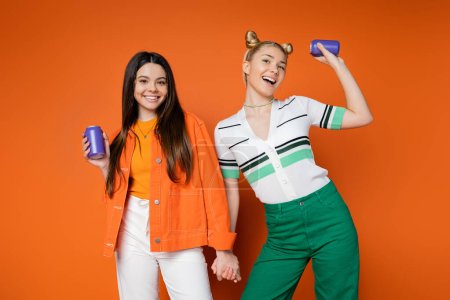 Fröhliche blonde und brünette Teenager-Freundinnen in lässigen Outfits mit Getränken in Blechdosen und Händchen haltend auf orangefarbenem Hintergrund, modische Mädchen mit Sinn für Stil