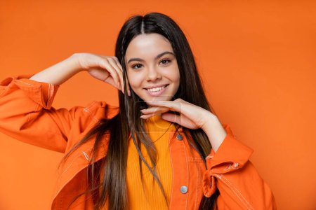 Portrait of trendy and brunette teen model in denim jacket touching face and looking at camera while standing and posing isolated on orange, cool and confident teenage girl