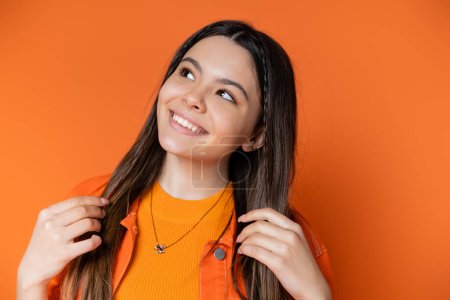 Porträt eines fröhlichen Teenie-Models in Jeansjacke, das brünettes Haar berührt und wegschaut, während es isoliert auf einem orangefarbenen, coolen und selbstbewussten Teenager-Mädchen posiert