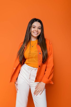 Fröhlich und brünett Teenager-Mädchen mit hellem Make-up trägt trendiges Outfit, während stehend und posiert auf orangefarbenem Hintergrund, cool und selbstbewusst Teenager-Mädchen