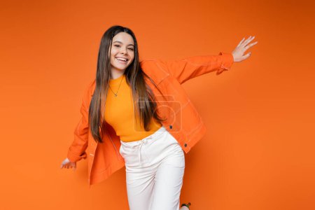 Photo for Fashionable and cheerful brunette teenage girl in denim jacket and pants looking at camera while posing isolated on orange, cool and confident teenage girl, gen z fashion - Royalty Free Image