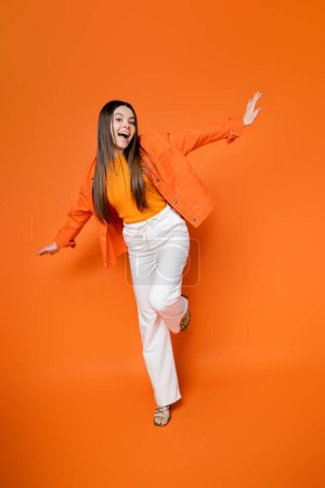 Full length of excited and trendy brunette teen model in denim jacket and pants posing and looking at camera while standing on orange background, cool and confident teenage girl
