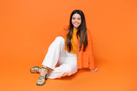 Jeune fille brune à la mode et souriante en talons et veste en denim regardant la caméra tout en étant assis et posant sur fond orange, adolescente cool et confiante, gen z fashion