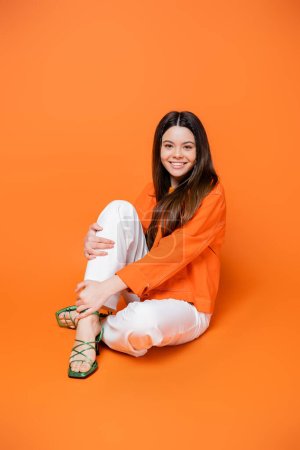 Full length of positive brunette teenager in heels and denim jacket in stylish outfit touching leg while sitting and posing on orange background, cool and confident teenage girl, gen z fashion