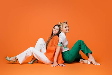 Full length of fashionable teenage girlfriends in casual outfits and heels smiling at camera while sitting back to back on orange background, cool and confident teenage girls