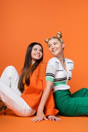 Fröhliche und trendige blonde und brünette Teenager-Freundinnen mit kühnem Make-up, die lächelnd Rücken an Rücken sitzen und einander auf orangefarbenem Hintergrund betrachten, coole und selbstbewusste Teenager-Mädchen