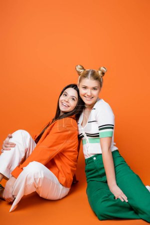 Photo for Positive and stylish blonde teenage model in casual outfit smiling at camera while sitting next to brunette girlfriend on orange background, cool and confident teenage girls - Royalty Free Image