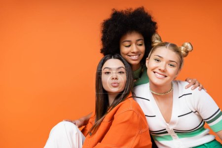 Fröhliche afrikanisch-amerikanische Teenager-Mädchen mit hellem Make-up umarmt stilvolle blonde und brünette Freundinnen, während sie zusammen isoliert auf orangefarbenen, coolen und selbstbewussten Teenager-Mädchen posieren