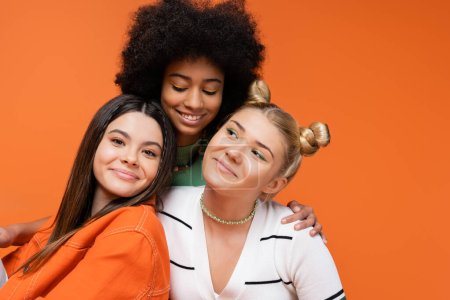 Lächelndes afrikanisch-amerikanisches Teenie-Model mit hellem Make-up, das modische Freundinnen umarmt und isoliert auf orangefarbenen, coolen und selbstbewussten Teenagermädchen posiert, diverse Rassen 