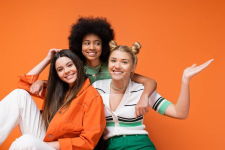 Lächelndes afrikanisch-amerikanisches Teenie-Mädchen mit hellem Make-up umarmt trendige Freundinnen, die in die Kamera schauen, während sie isoliert auf orangefarbenen, coolen und selbstbewussten multikulturellen Teenagermädchen sitzen